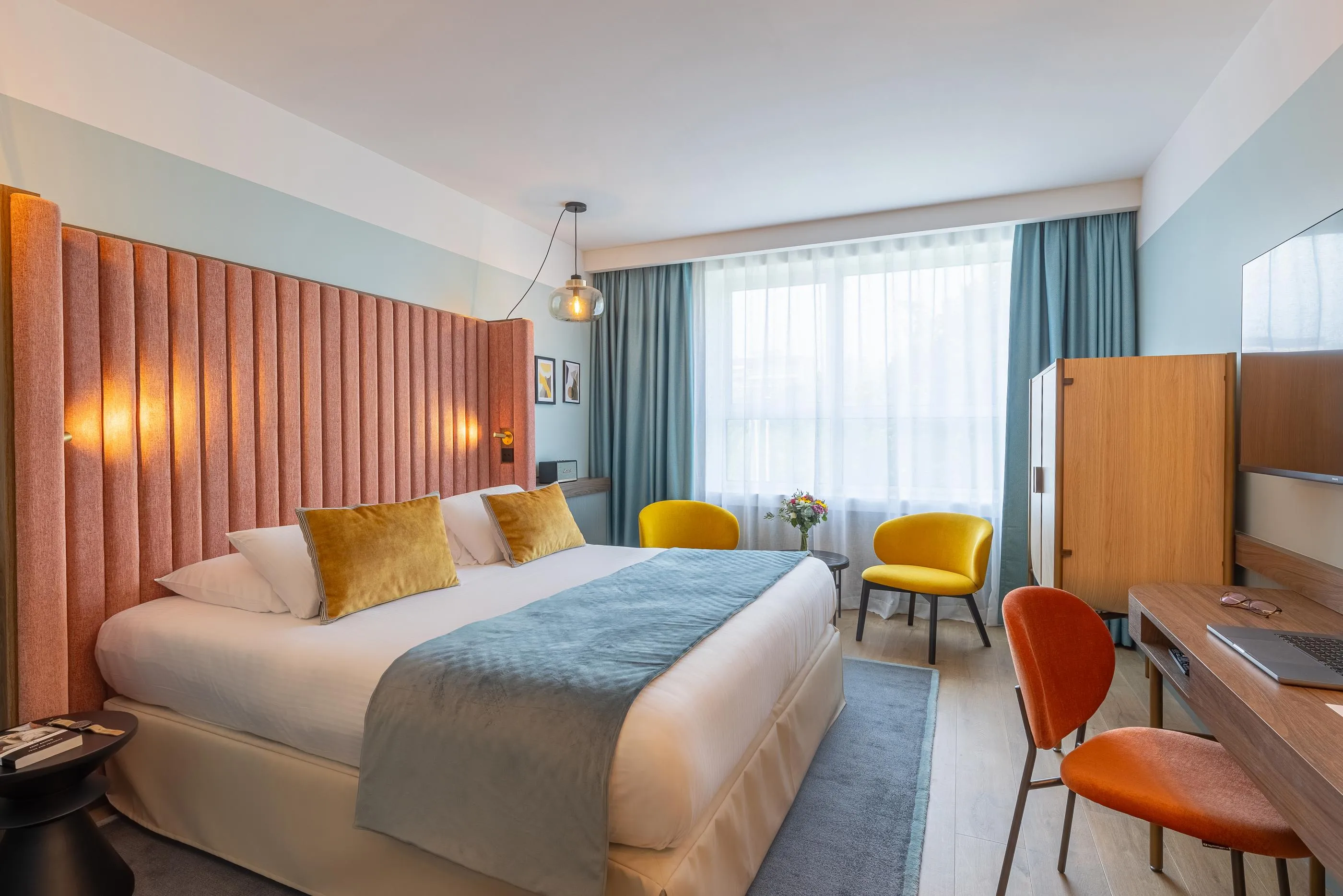 Chambre d'hôtel avec un lit double, deux chaises colorées, un bureau et de grandes fenêtres avec des rideaux