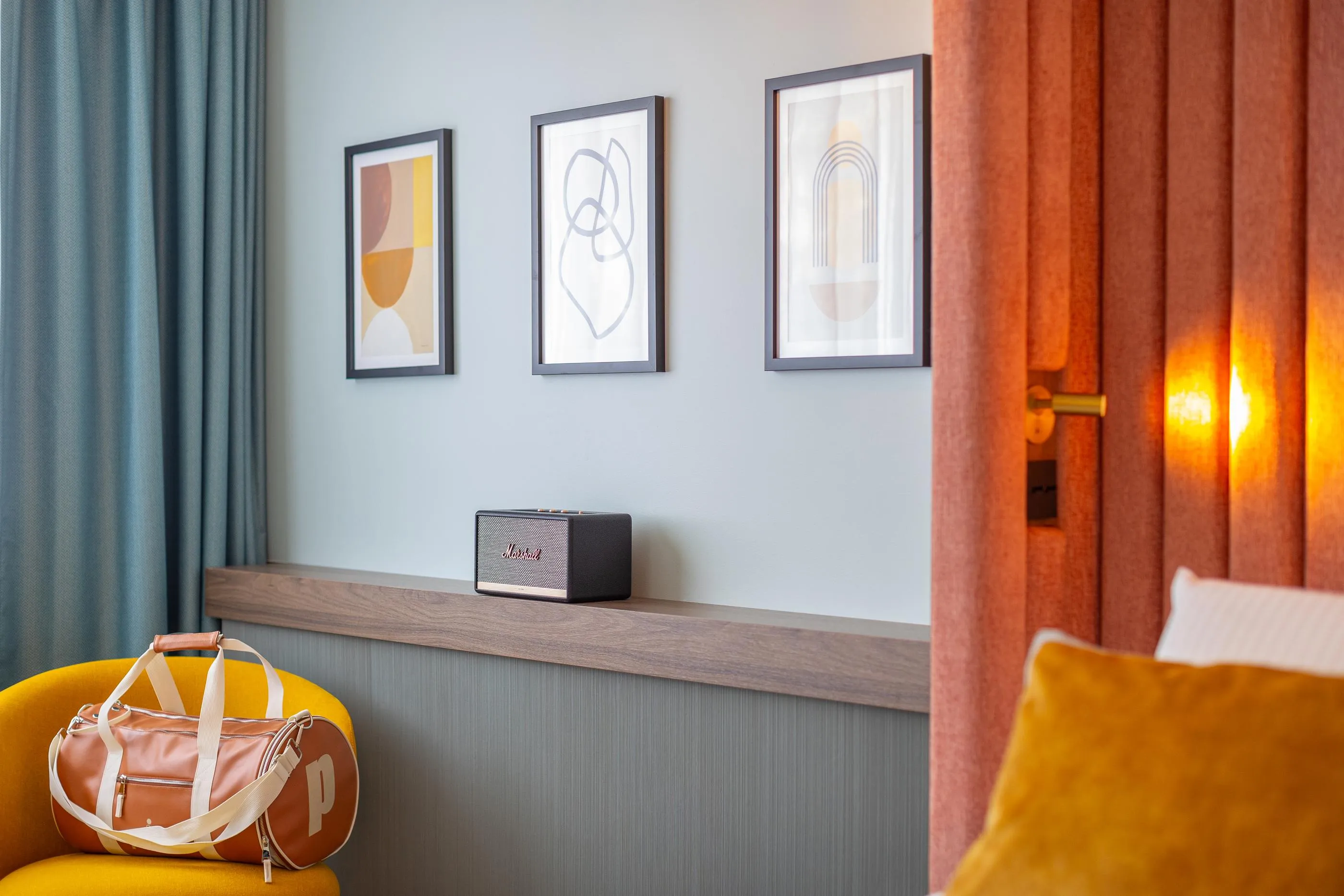 Un salon avec des œuvres d'art encadrées sur le mur, un haut-parleur sur une étagère, un sac en cuir sur un fauteuil jaune et une porte entrouverte avec de la lumière filtrant à travers les rideaux