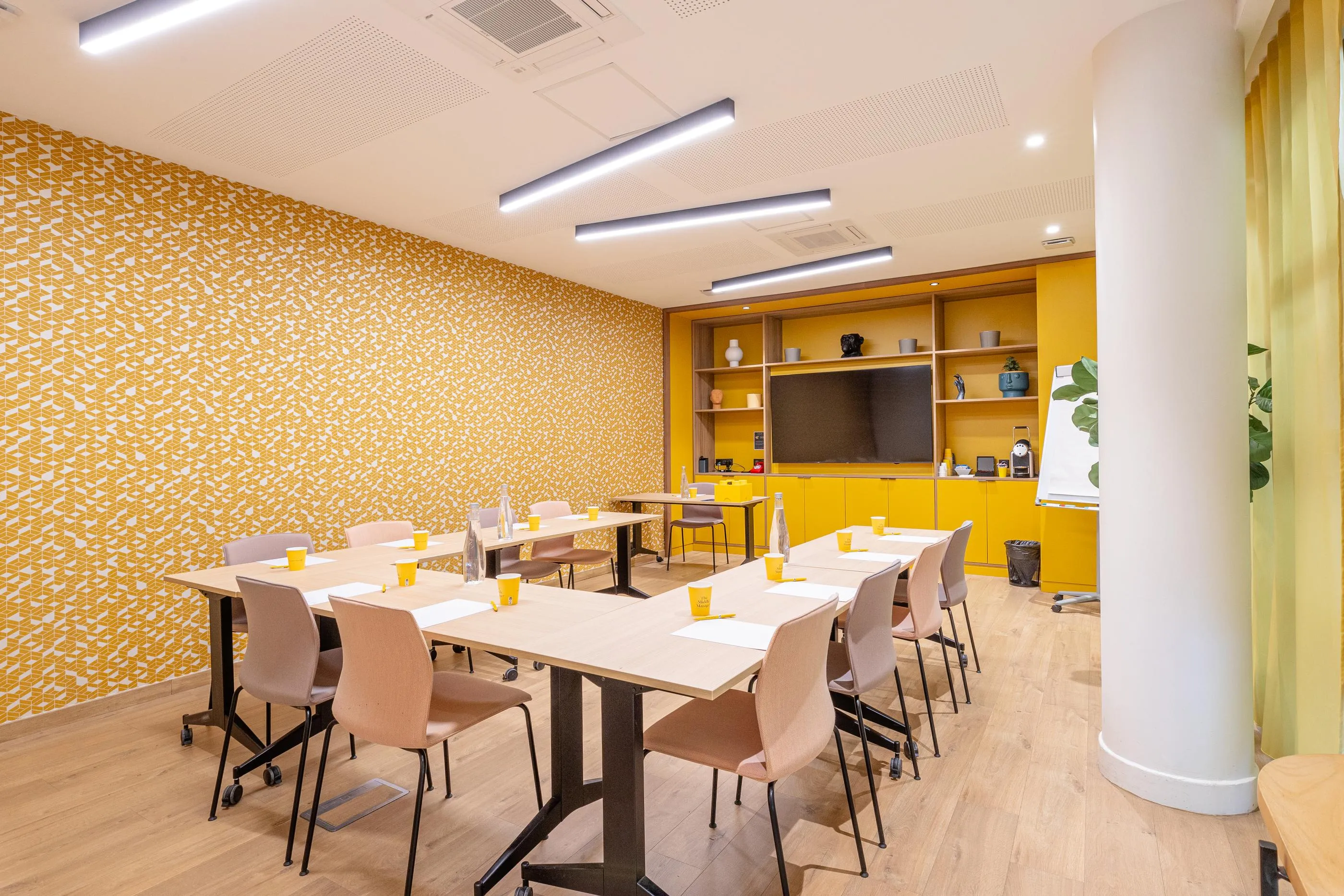 Salle de réunion avec tables, chaises et écran, murs jaunes à motifs géométriques et éclairage moderne