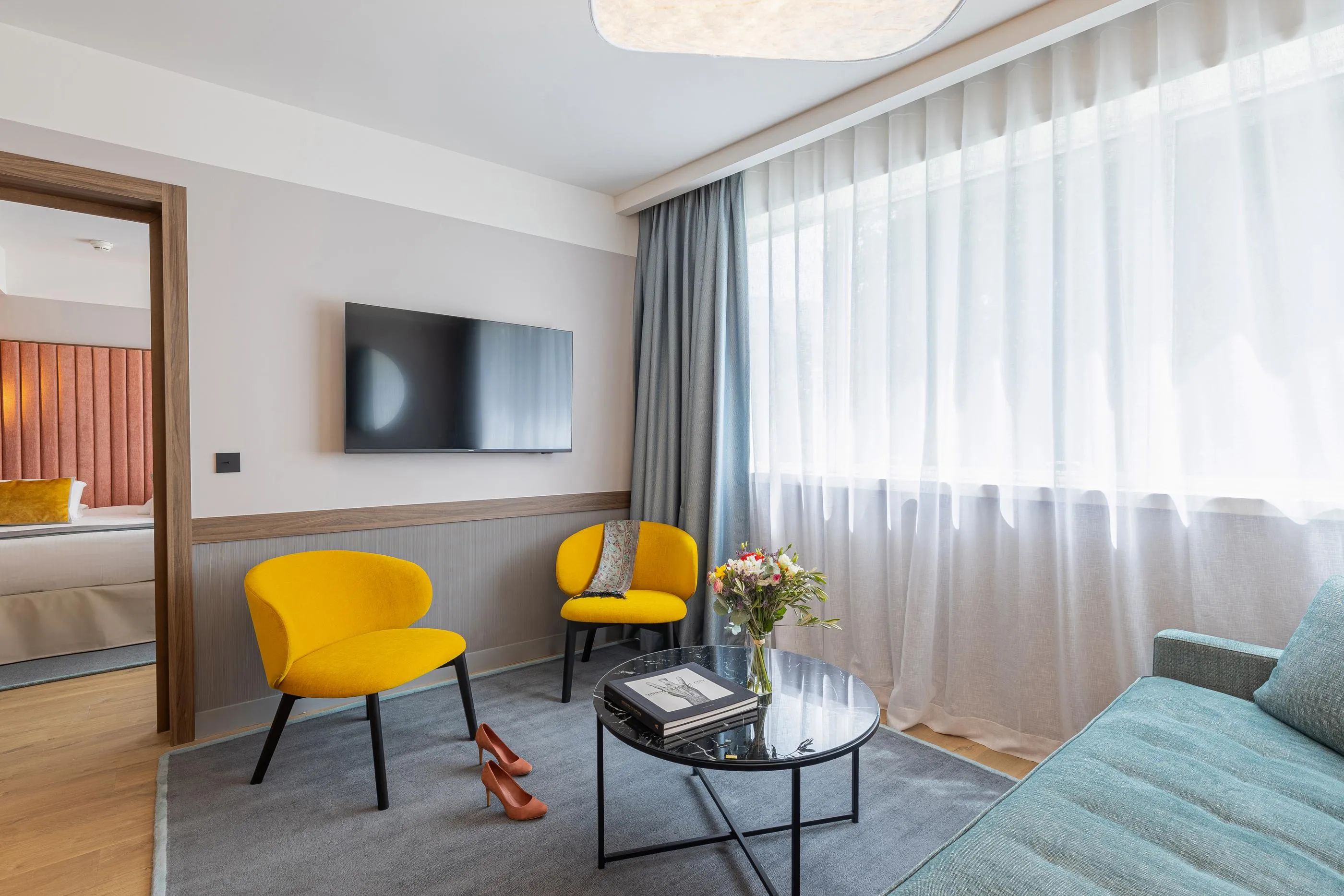 Salon d'hôtel avec deux fauteuils jaunes, une table basse, des magazines, un vase de fleurs et une télévision murale
