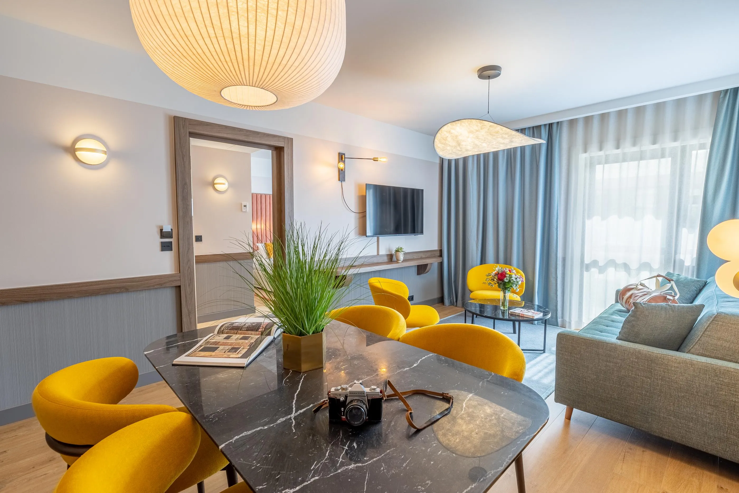 Salon moderne avec table en marbre noir, chaises jaunes, canapé gris, plantes vertes et télévision murale