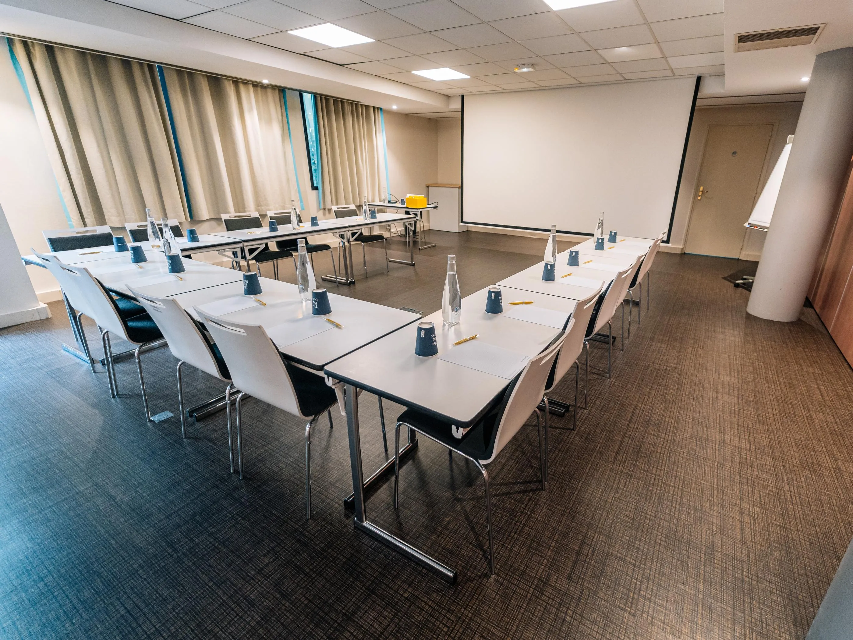 Salle de réunion avec plusieurs tables disposées en U, chacune équipée de bouteilles d'eau, de gobelets et de stylos