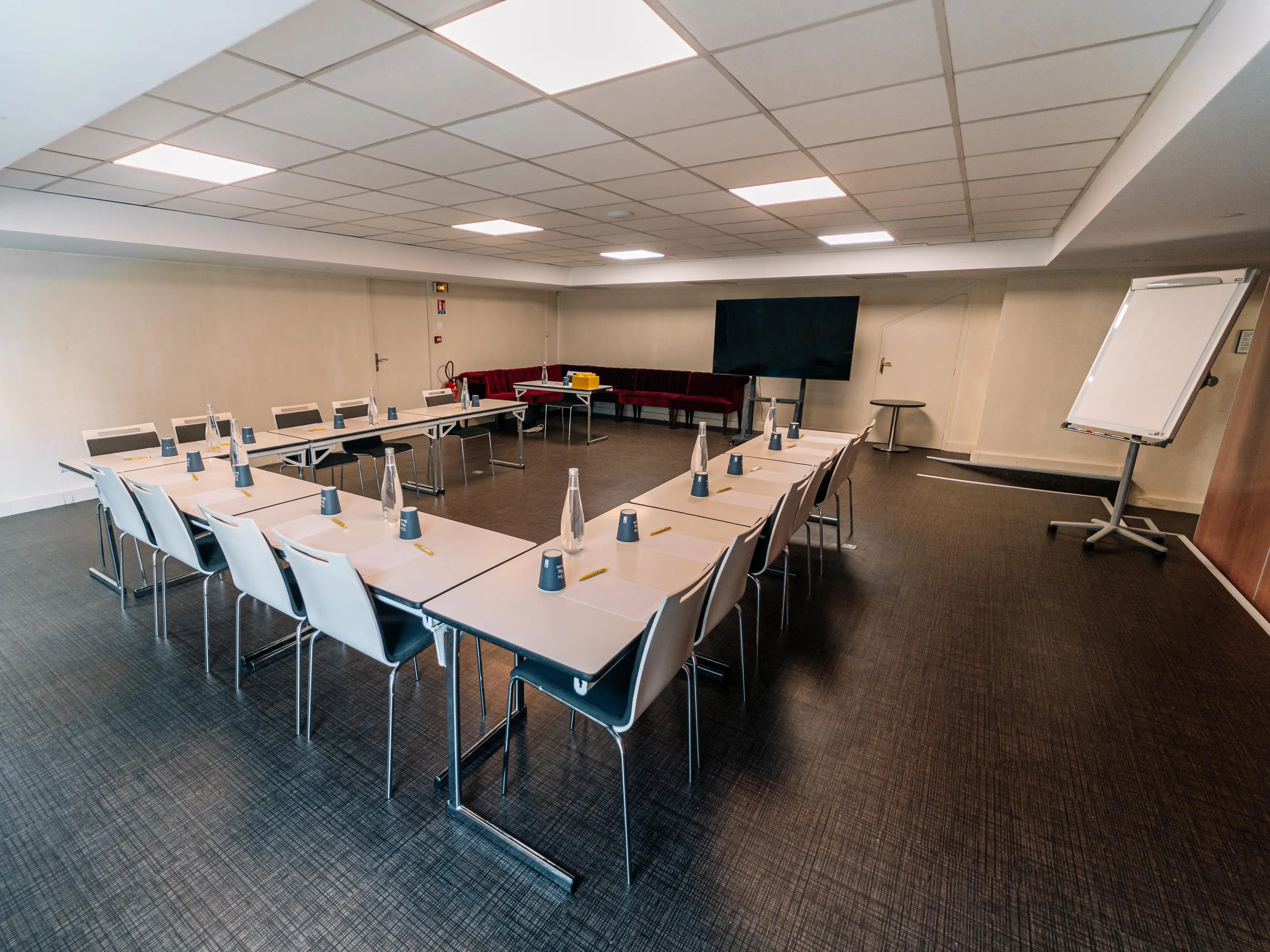 Salle de réunion avec tables disposées en U, chaises, écran, tableau blanc et rangée de sièges rouges