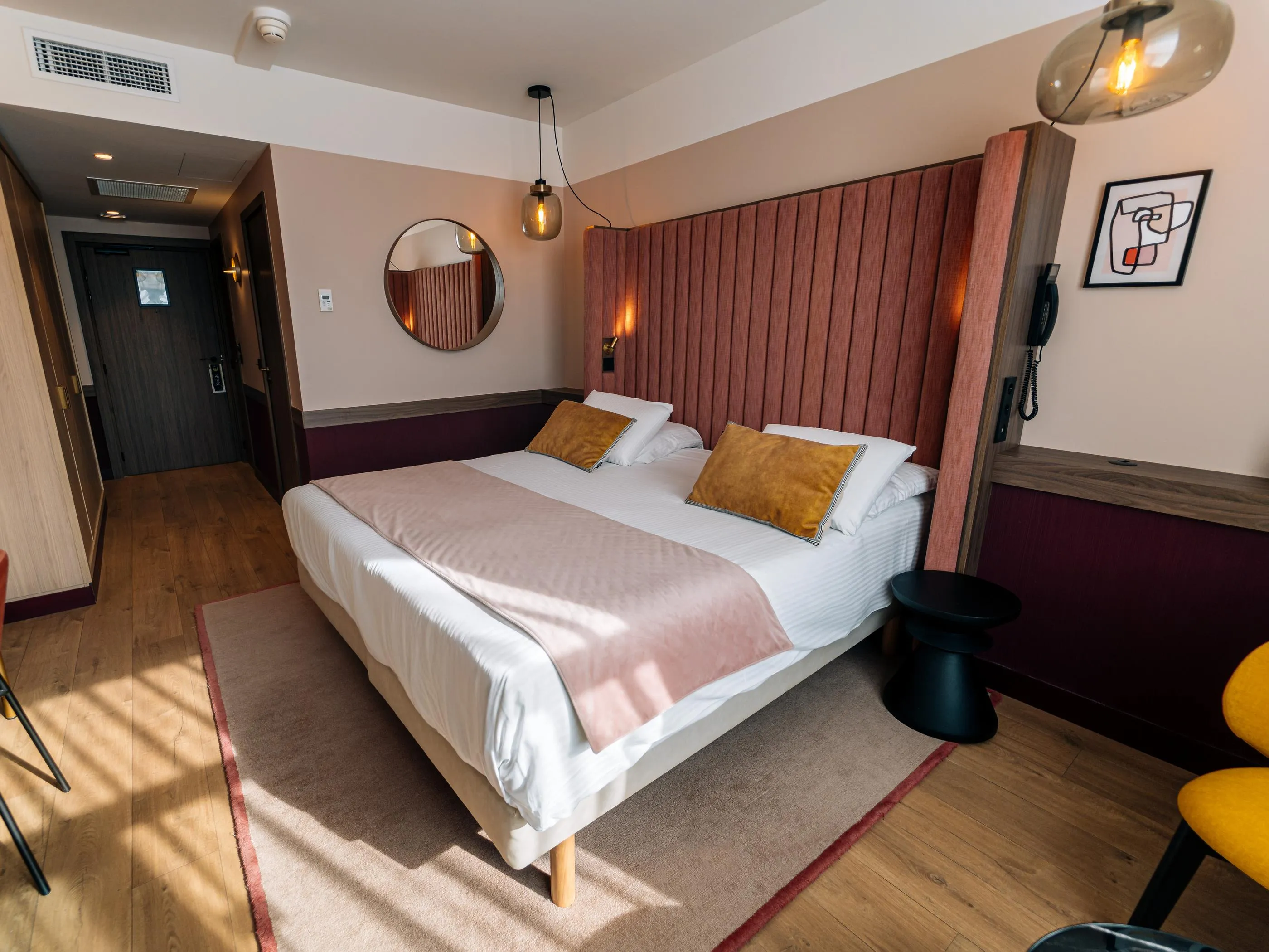 Chambre avec un lit double, des coussins dorés, une tête de lit en bois, une lampe murale et un miroir rond