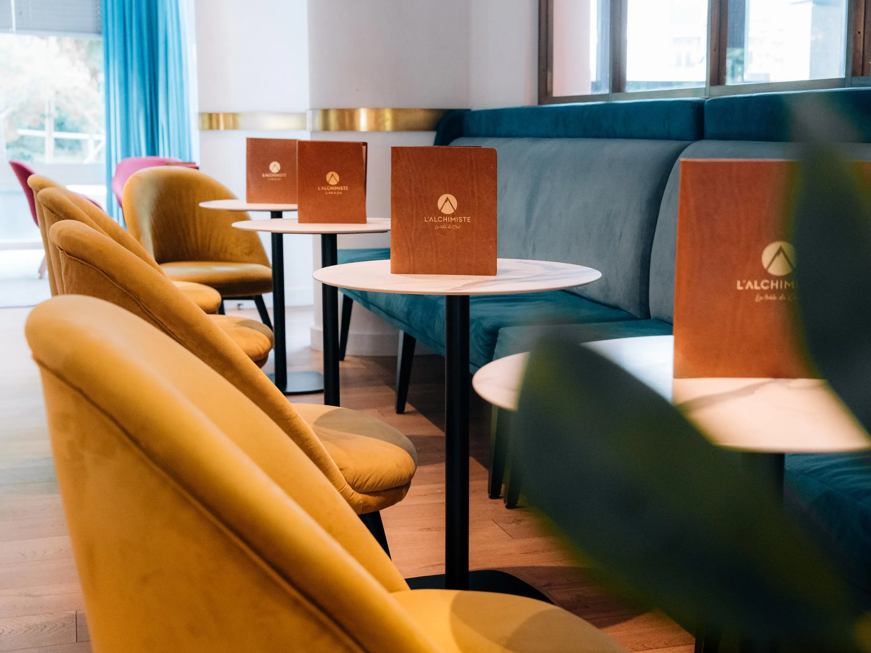 Tables avec menus "L'Alchimiste" dans un espace intérieur avec des sièges en velours jaune et des banquettes vertes