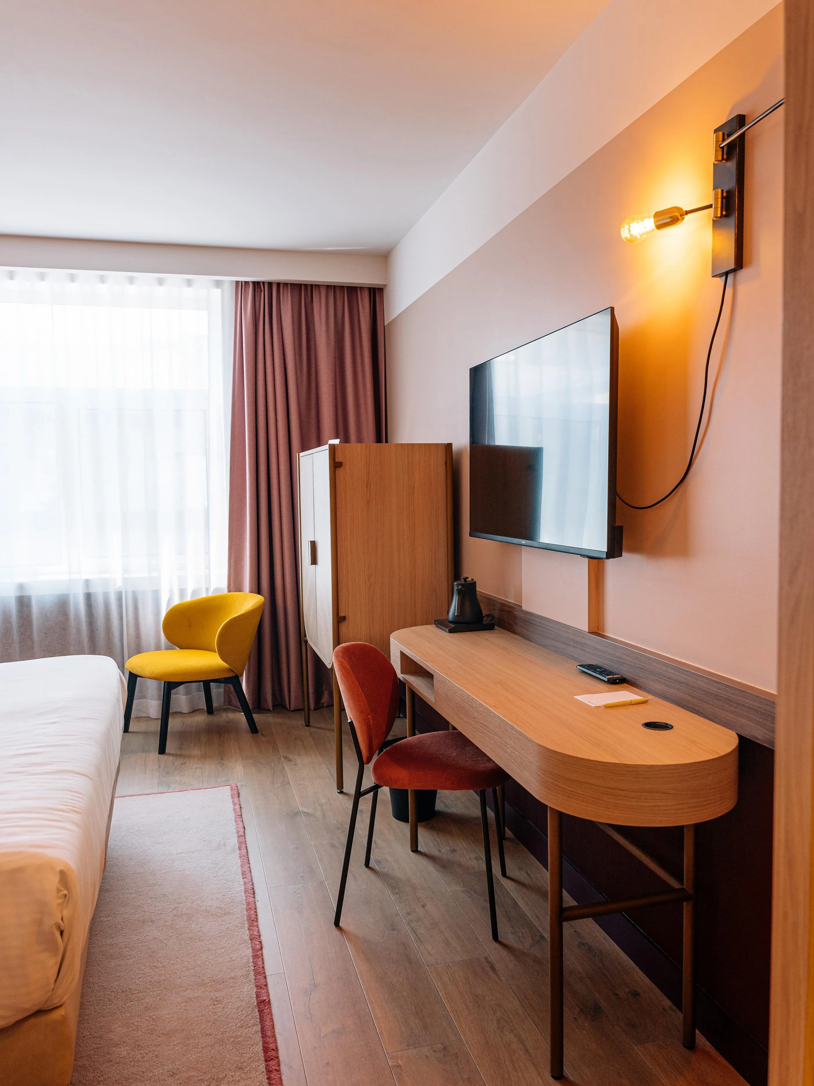 Chambre avec un lit, une table de travail, une télévision, un fauteuil jaune et une fenêtre avec des rideaux
