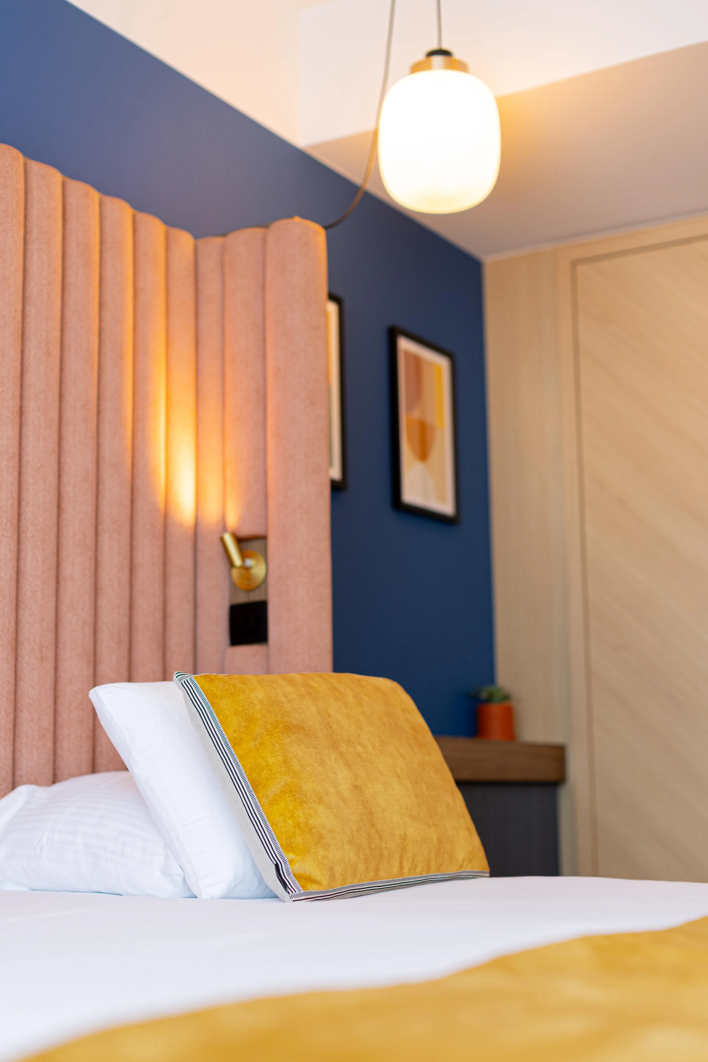 Lit avec une couverture blanche et un coussin jaune, une lampe murale allumée et une suspension au plafond