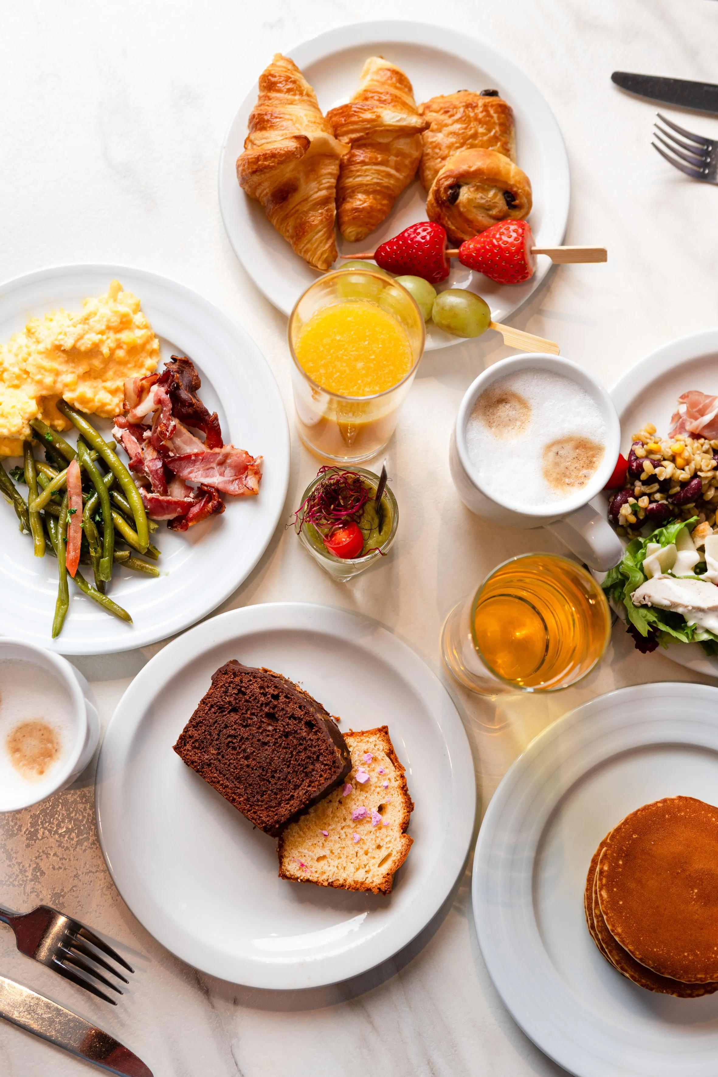 Un assortiment de plats variés incluant des croissants, des œufs brouillés, des asperges, du bacon, des pancakes, des fruits et des boissons sur une table