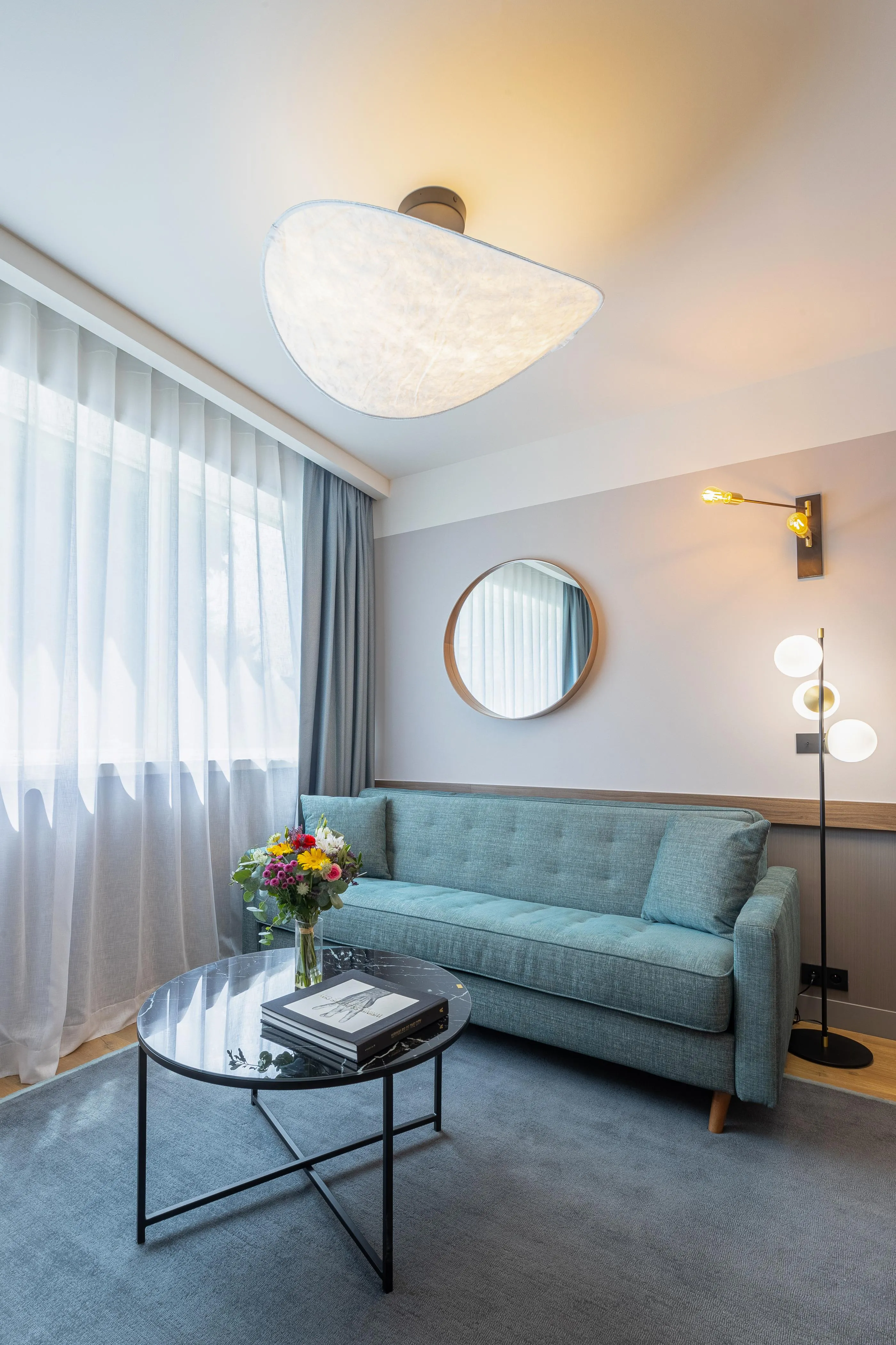 Un canapé vert, une table basse avec des livres et des fleurs, une lampe à trois globes et un miroir rond dans un salon moderne