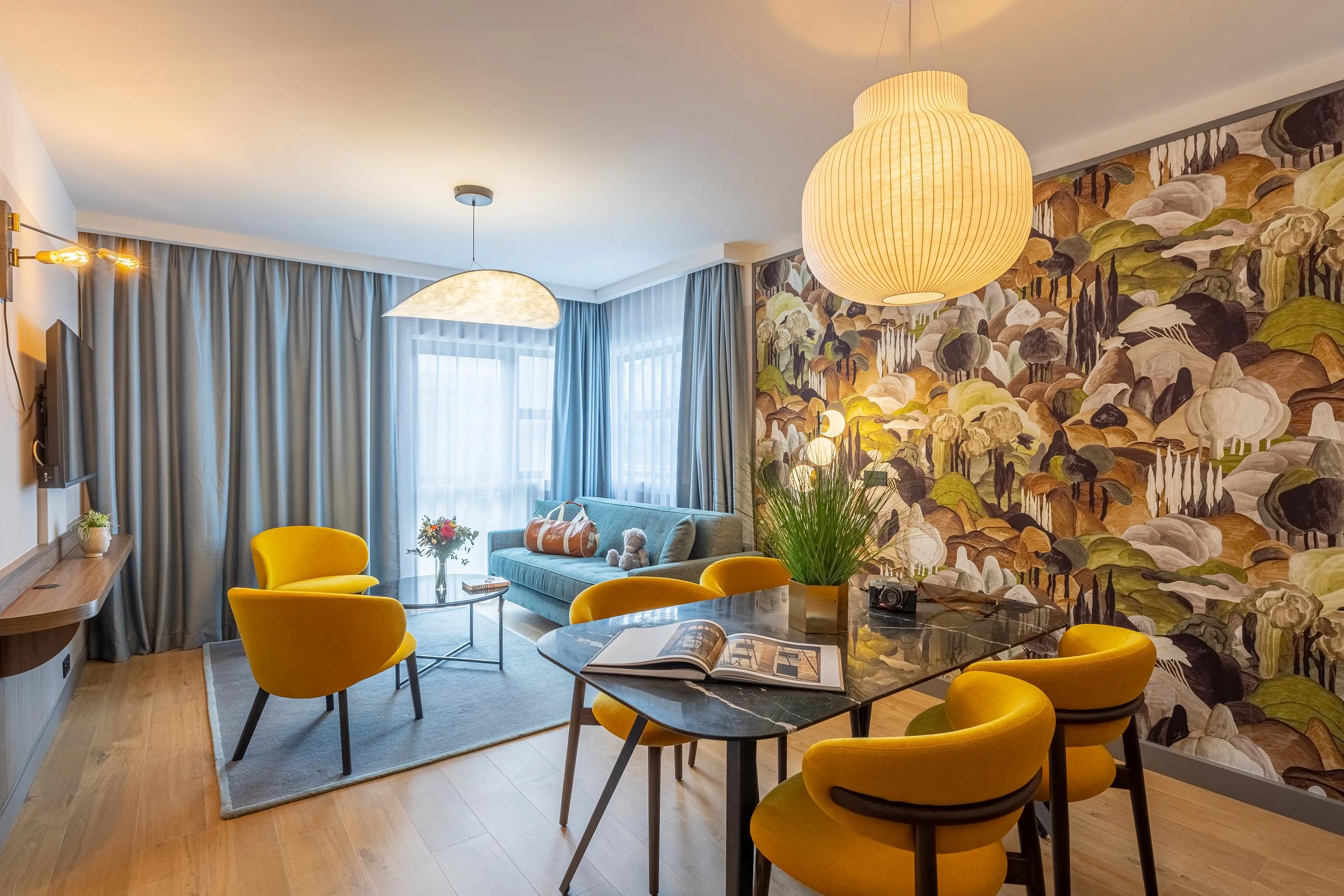 Salon moderne avec canapé bleu, chaises jaunes, table en verre et mur décoré de motifs floraux et champignons