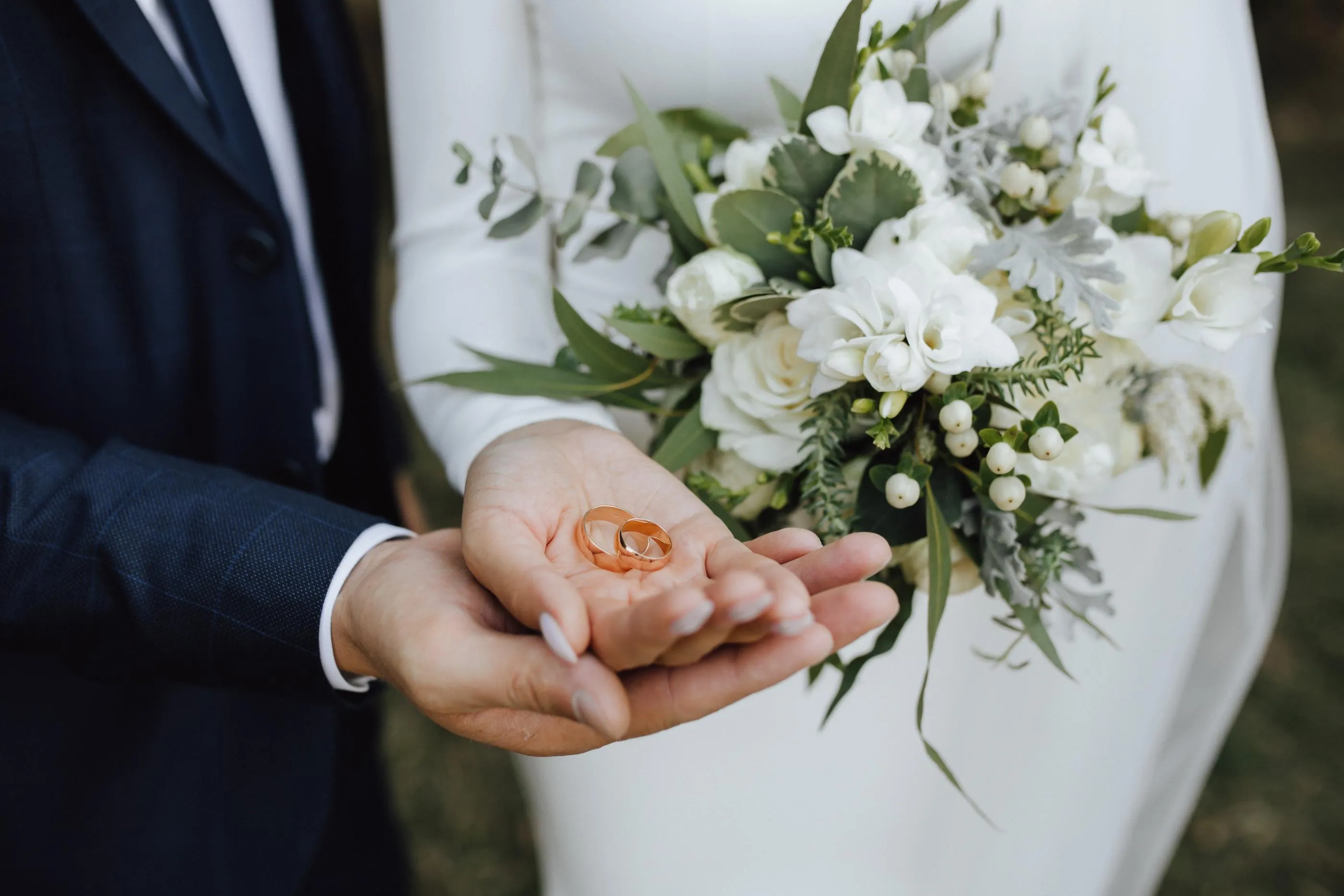 Deux mains tiennent des alliances au-dessus d'un bouquet de fleurs blanches et vertes