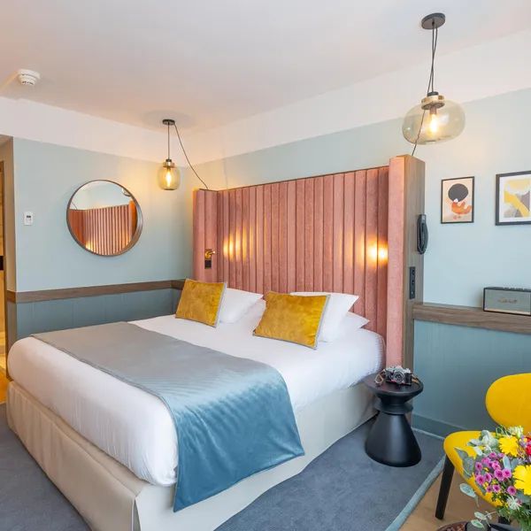 Chambre avec un lit double, une tête de lit en tissu, des coussins jaunes, une table de chevet, un fauteuil jaune et des fleurs sur la table