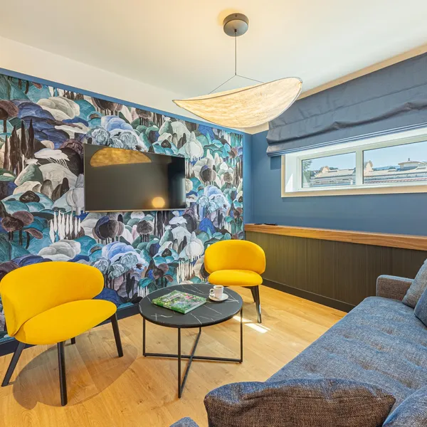 Salon avec canapé bleu, deux fauteuils jaunes, une table basse, un mur décoré d'un grand motif floral et une télévision