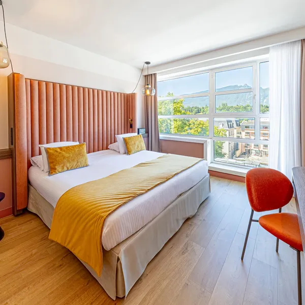 Chambre d'hôtel avec un lit double, une table de travail, une fenêtre offrant une vue sur des montagnes et des bâtiments, et des luminaires suspendus