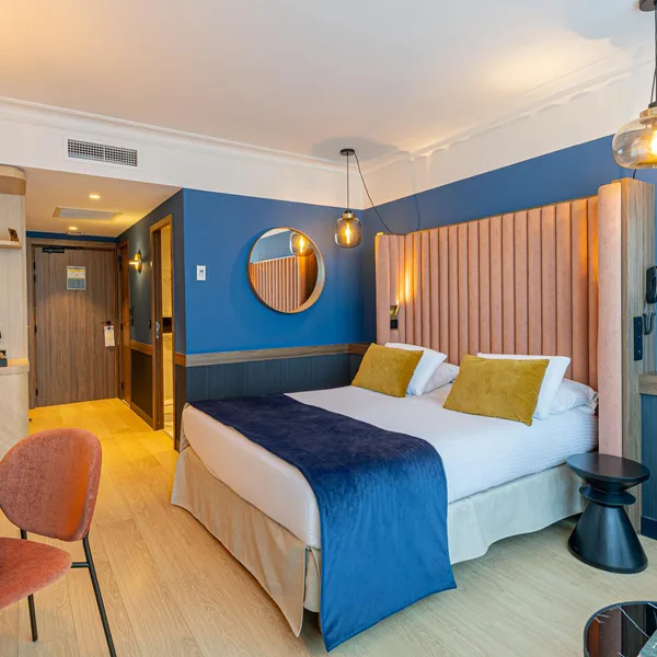 Chambre d'hôtel avec lit double, table de travail, deux chaises colorées et un coin salon, murs peints en bleu et décorations murales