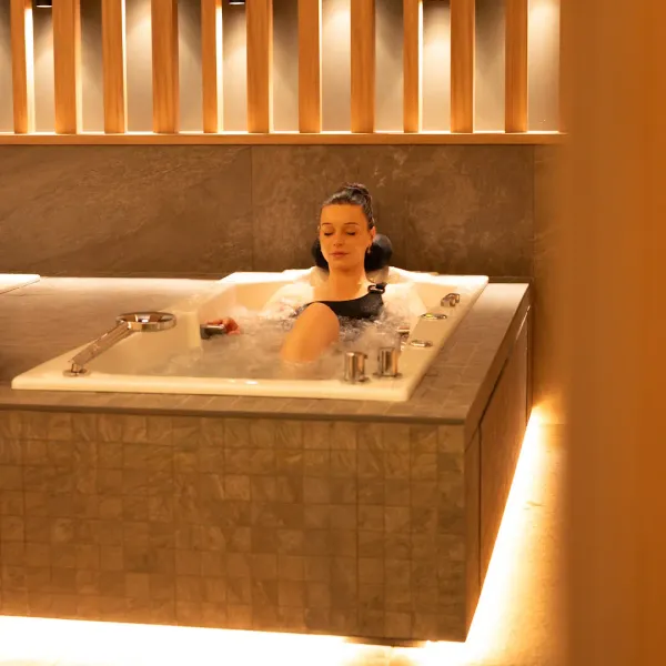 Une femme est allongée dans un bain à remous entouré de carreaux en bois clair et éclairé par une lumière dorée