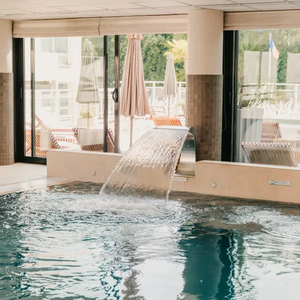 Piscine intérieure avec un petit bassin et une cascade, entourée de grandes baies vitrées ouvrant sur une terrasse avec des chaises longues