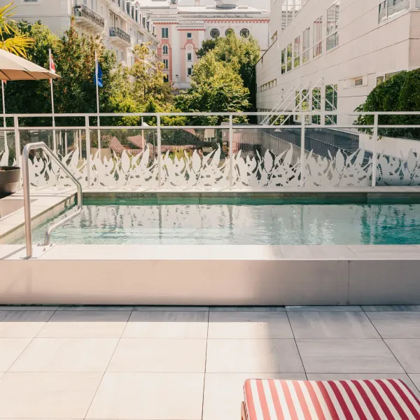 Piscine rectangulaire avec escaliers et main courante, entourée d'un sol en carreaux clairs, de transats et de plantes en pot, avec un bâtiment en arrière-plan