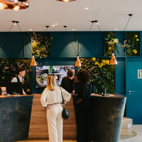 Trois personnes discutent devant un comptoir dans un espace intérieur moderne aux murs verts décorés de plantes, avec des lampes suspendues au plafond