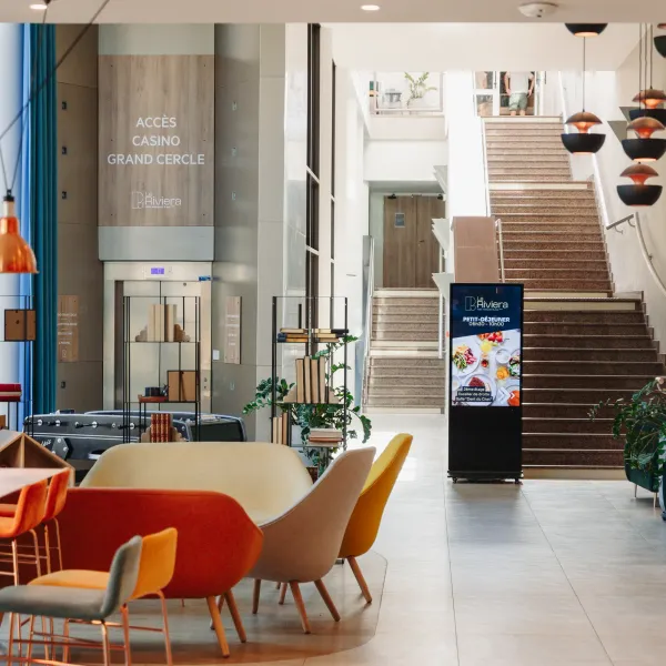 Hall d'entrée moderne avec escalier, sièges colorés, plantes et panneaux d'affichage indiquant l'accès au casino et les horaires du petit-déjeuner