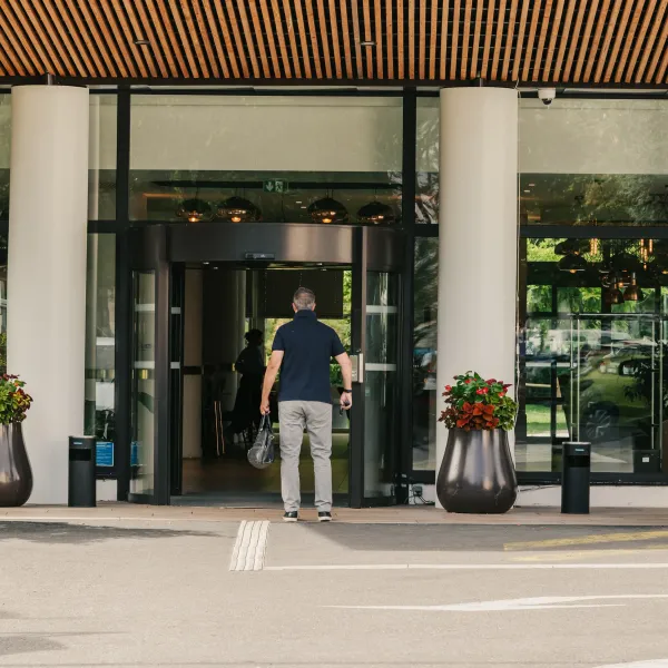 Un homme se tient devant l'entrée d'un bâtiment moderne avec des portes vitrées, des plantes en pot et un trottoir