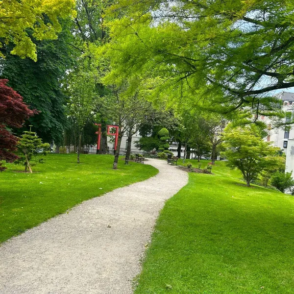 Un chemin de gravier sinueux traverse un parc arboré avec des bancs et des arbres de différentes couleurs