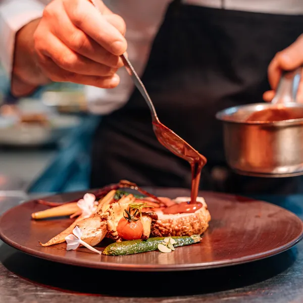 Un chef de cuisine verse une sauce sur un plat composé de viande, légumes et garnitures sur une assiette ronde