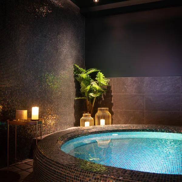 Jacuzzi intérieur avec éclairage bleu, entouré de mosaïques et de plantes, avec des bougies et une lampe à proximité