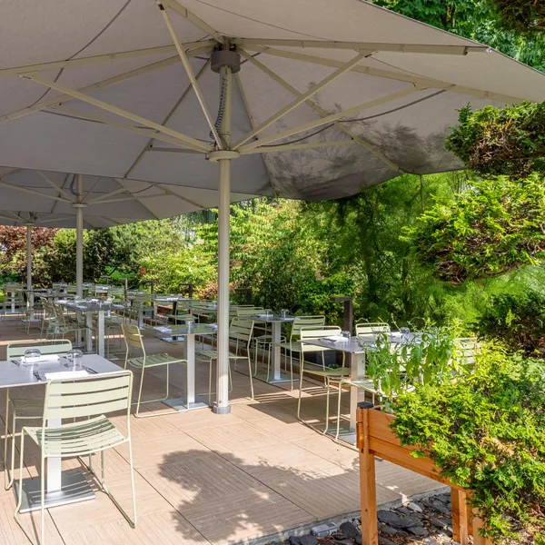Terrasse de restaurant extérieure avec tables et chaises sous un grand parasol, entourée de verdure