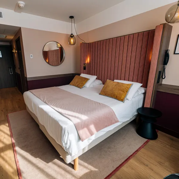 Chambre avec un lit double, des coussins dorés, une tête de lit en bois, une lampe murale et un miroir rond