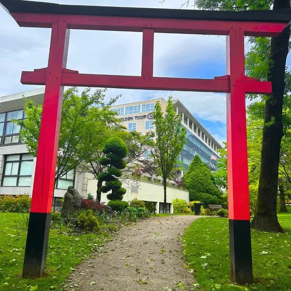 Un torii rouge se dresse dans un jardin avec des arbres et des bâtiments modernes en arrière-plan