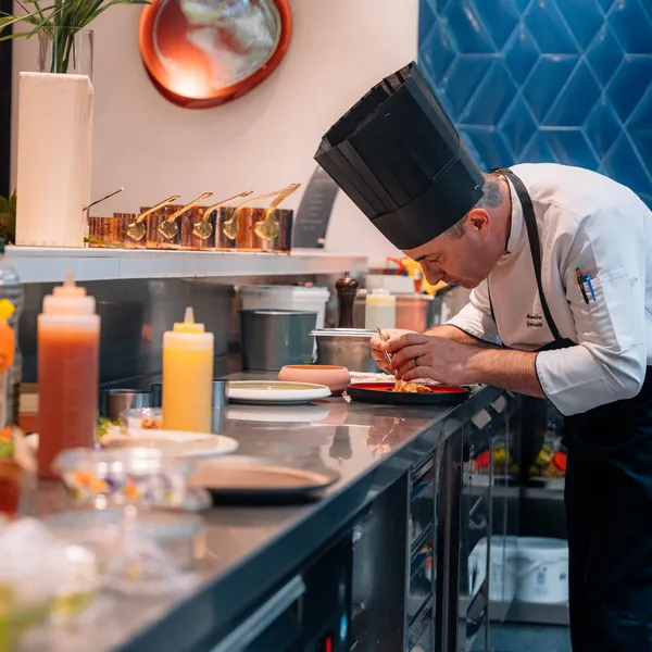 Un chef en toque blanche prépare une assiette dans une cuisine professionnelle avec divers ingrédients et ustensiles autour de lui