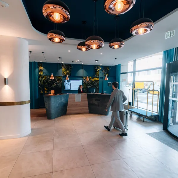 Un homme pousse un chariot à bagages vers un comptoir de réception d'hôtel moderne avec des luminaires suspendus et des plantes décoratives