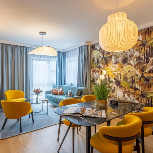 Salon moderne avec canapé bleu, chaises jaunes, table en verre et mur décoré de motifs floraux et champignons