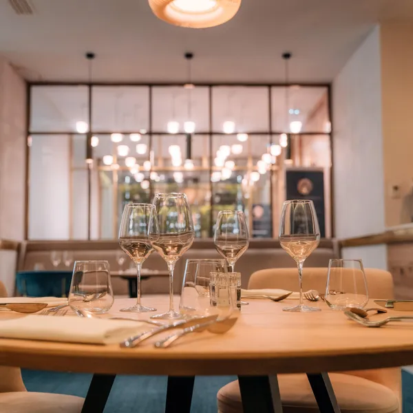 Table dressée avec verres à vin et couverts, vue depuis un angle, dans un restaurant aux lumières tamisées et aux grandes fenêtres