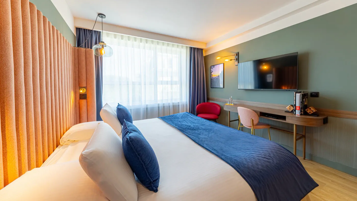Chambre d'hôtel avec un lit double, une télévision, un bureau, une chaise et une grande fenêtre avec des rideaux