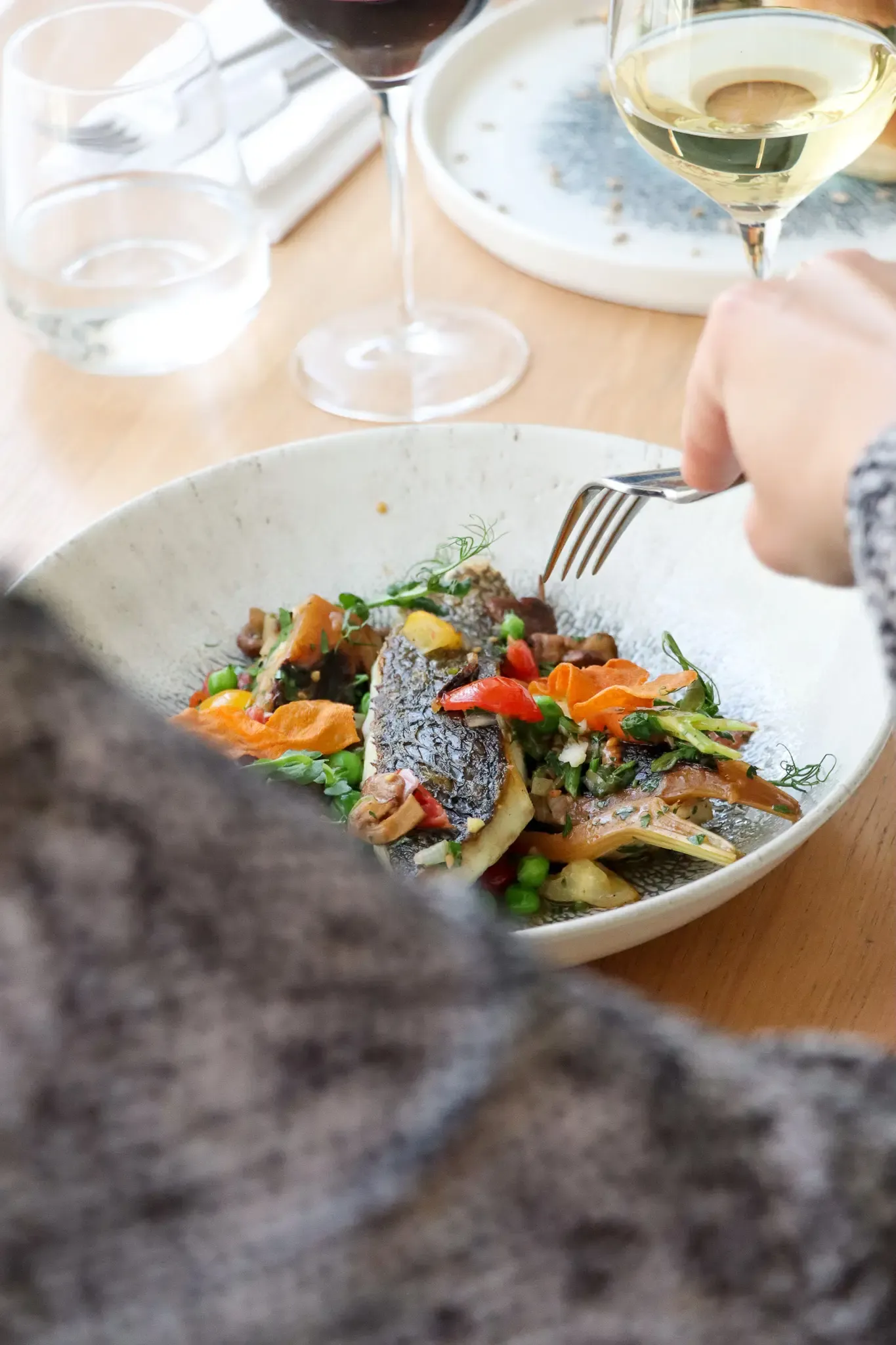 Un poisson grillé accompagné de légumes et de carottes, servi dans une assiette blanche, avec une main tenant une fourchette et des verres de vin à l'arrière-plan