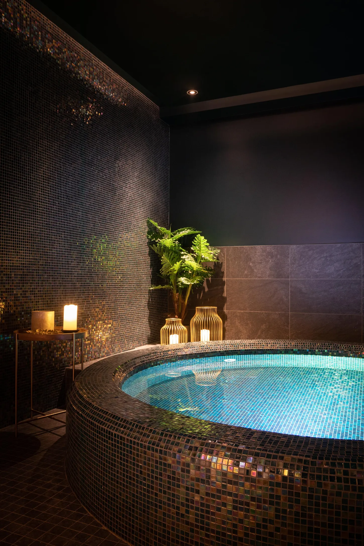 Innenjacuzzi mit blauer Beleuchtung, umgeben von Mosaiken und Pflanzen, mit Kerzen und einer Lampe in der Nähe