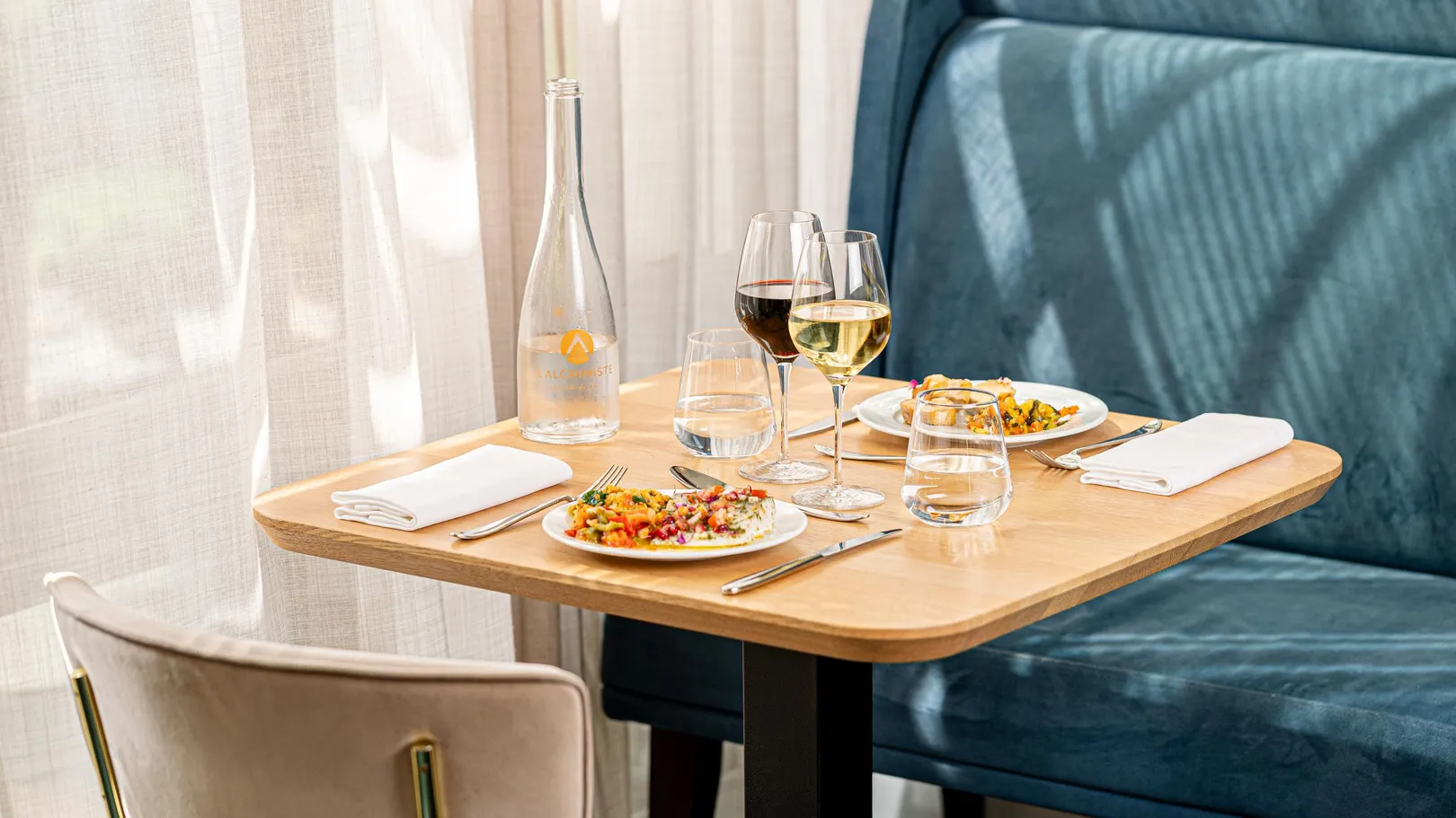 Table dressée avec deux assiettes de nourriture, deux verres de vin, une bouteille d'eau et des verres d'eau, près d'une fenêtre avec des rideaux blancs