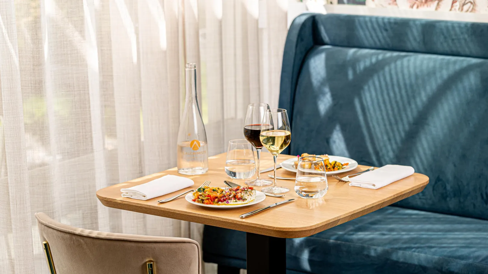 Table dressée avec deux assiettes de salade, deux verres de vin, deux verres d'eau, une bouteille d'eau et une nappe blanche sur un fond de banquette bleue et de rideaux blancs