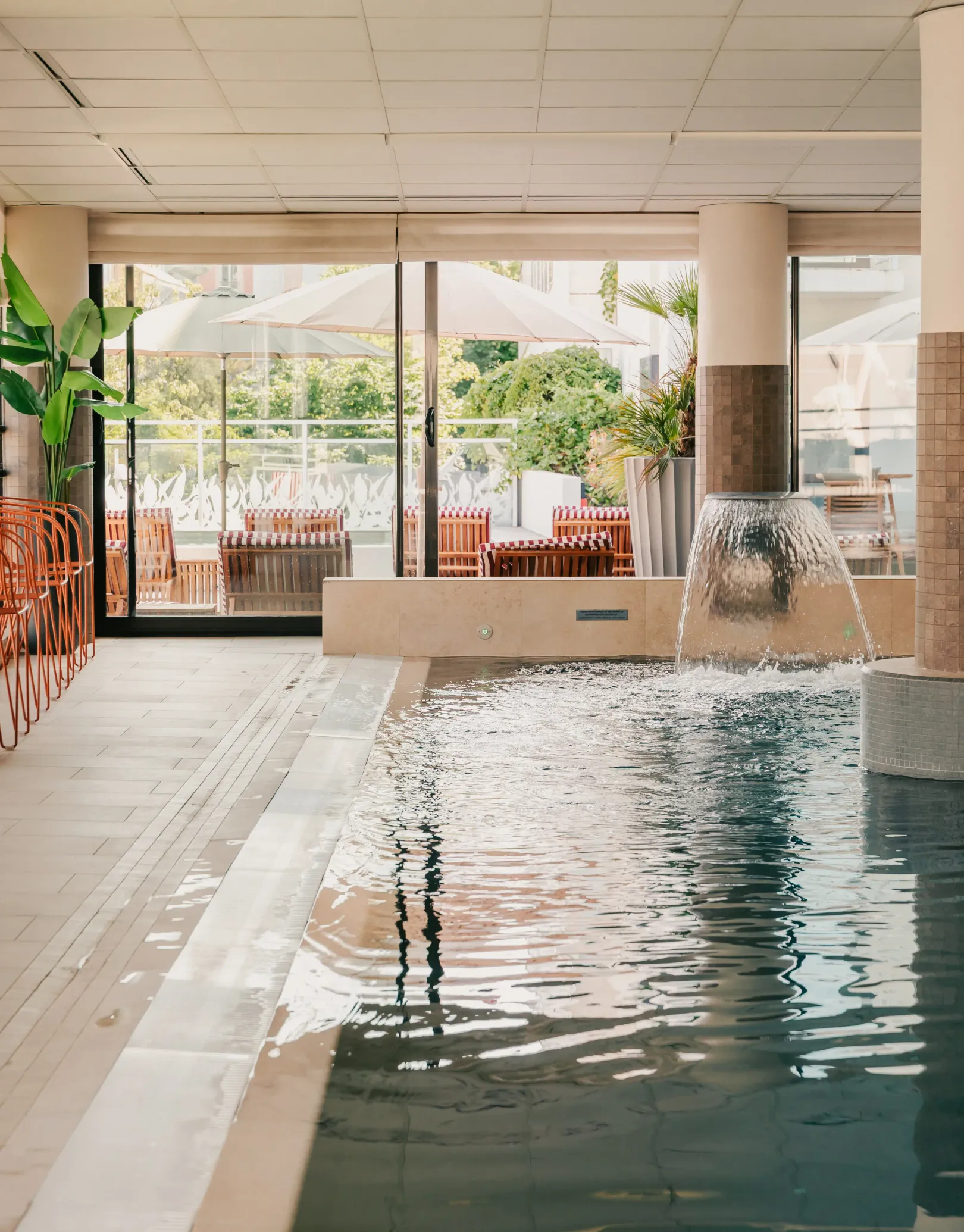 Piscine intérieure avec une fontaine et des tables en terrasse visibles à travers les baies vitrées