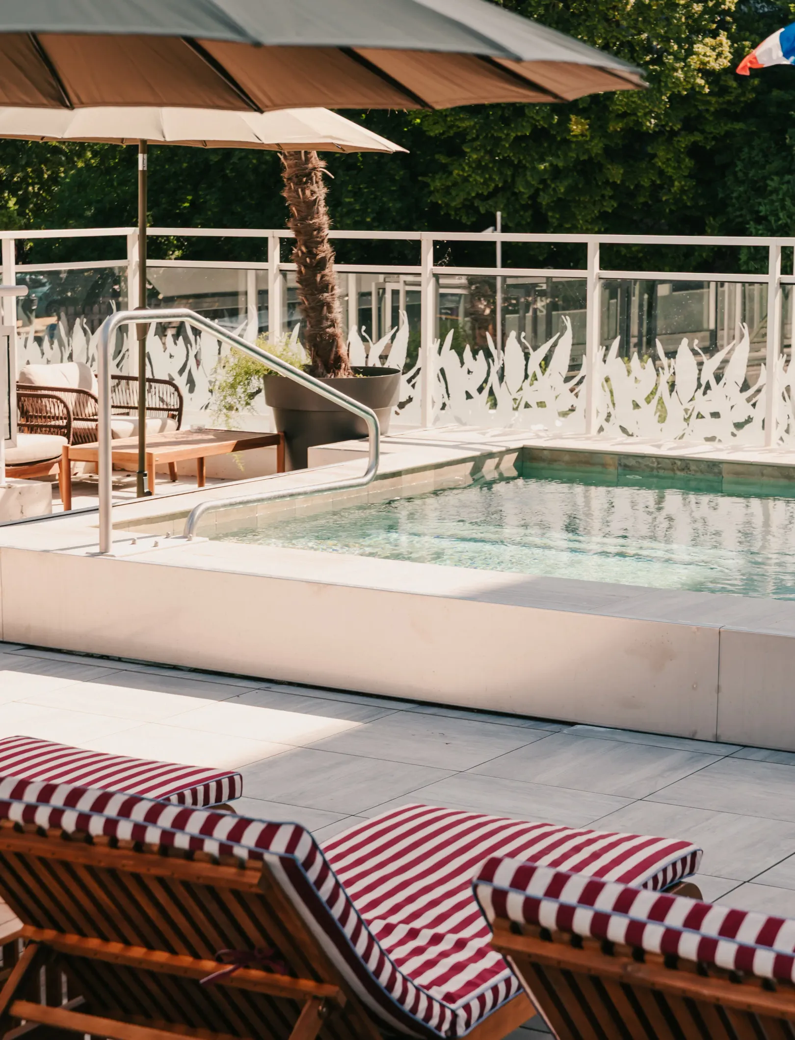 Schwimmbecken mit rot-weiß gestreiften Liegestühlen, Sonnenschirmen und Topfpalmen auf einer Terrasse mit einer Glasbrüstung