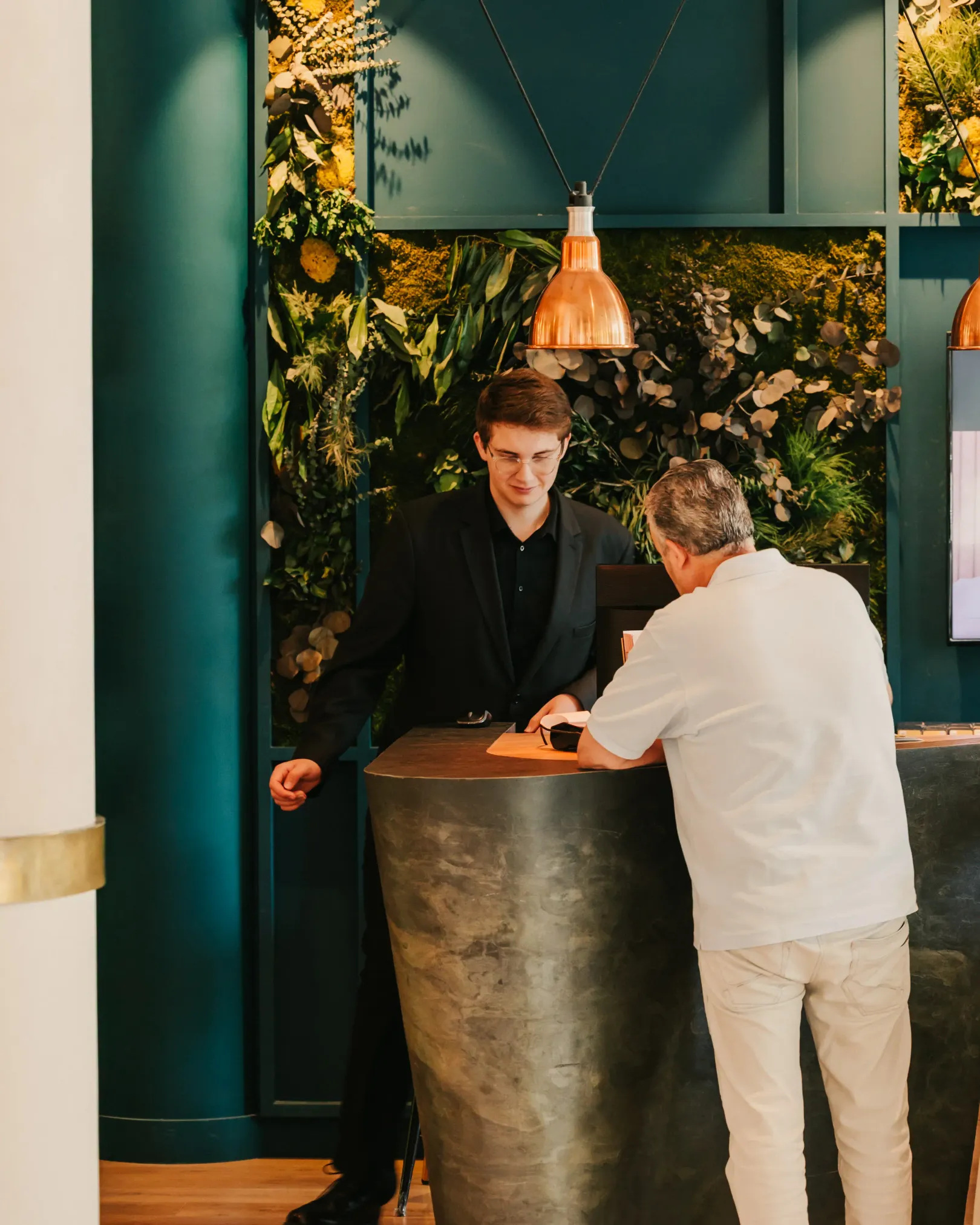 Deux hommes discutent à un comptoir devant un mur décoré de plantes vertes et de fleurs