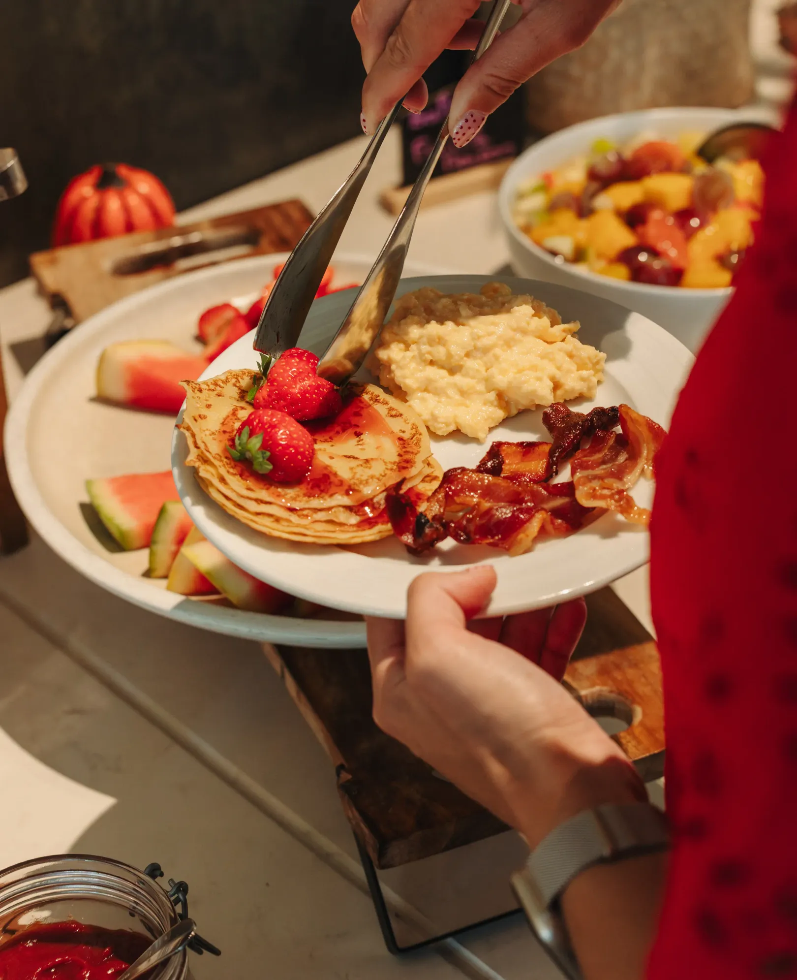 Une personne sert des pancakes garnis de fraises, de bacon et de purée de fruits sur une assiette, entourée de fruits et d'autres plats