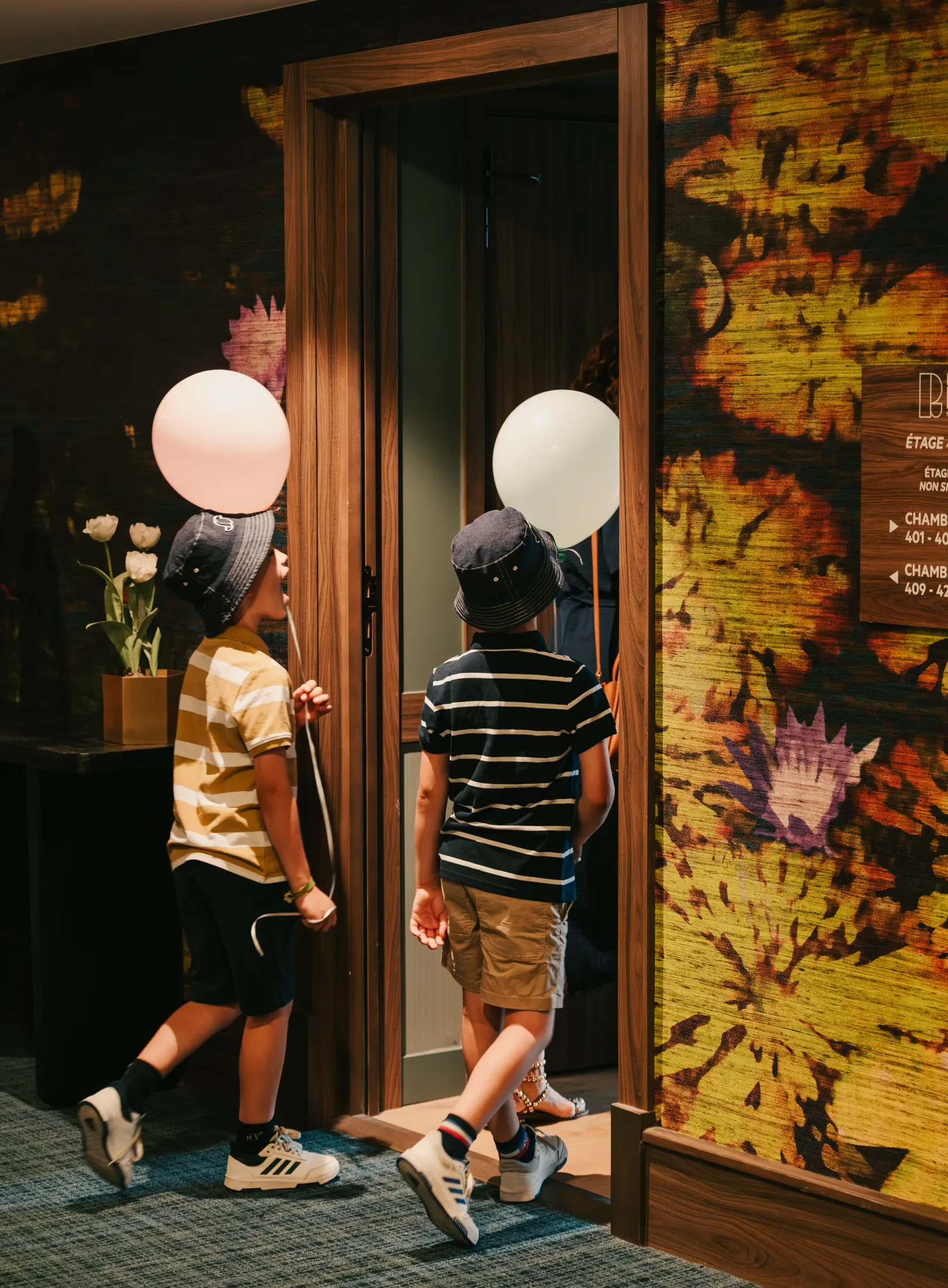 Deux enfants portant des chapeaux et des ballons blancs marchent devant une porte en bois avec un mur décoré de motifs colorés à côté