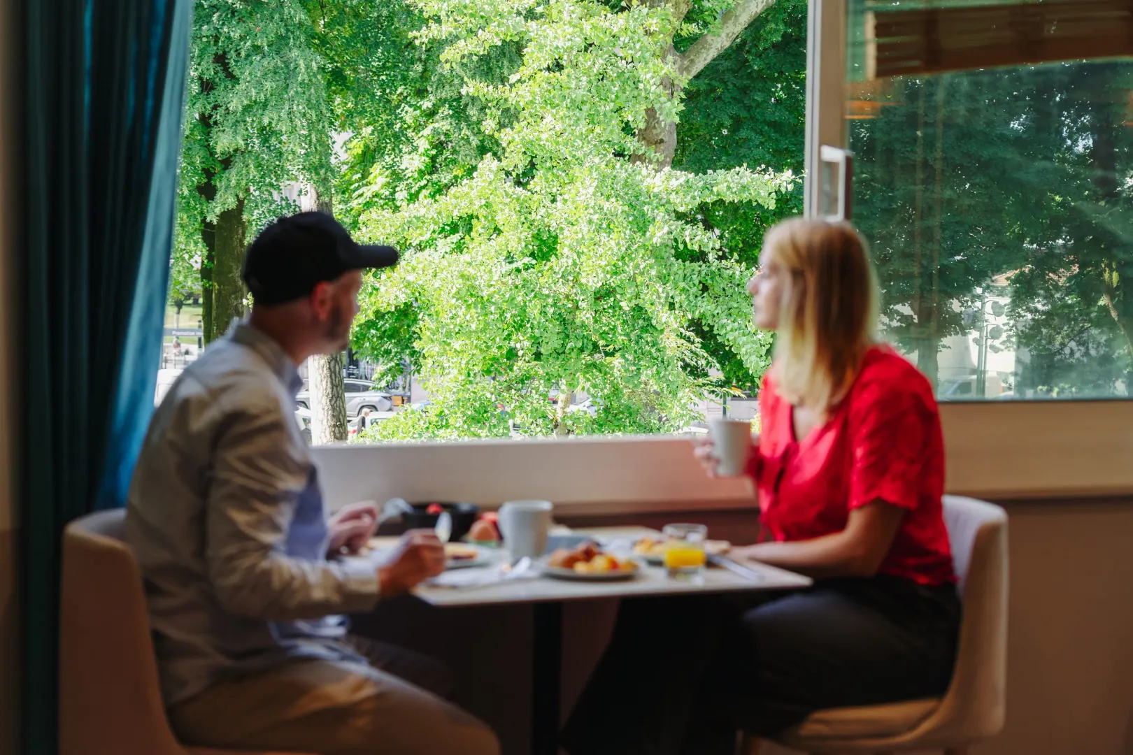 Deux personnes assises à une table avec de la nourriture et des boissons, discutant dans un cadre intérieur avec une grande fenêtre donnant sur des arbres en fleurs