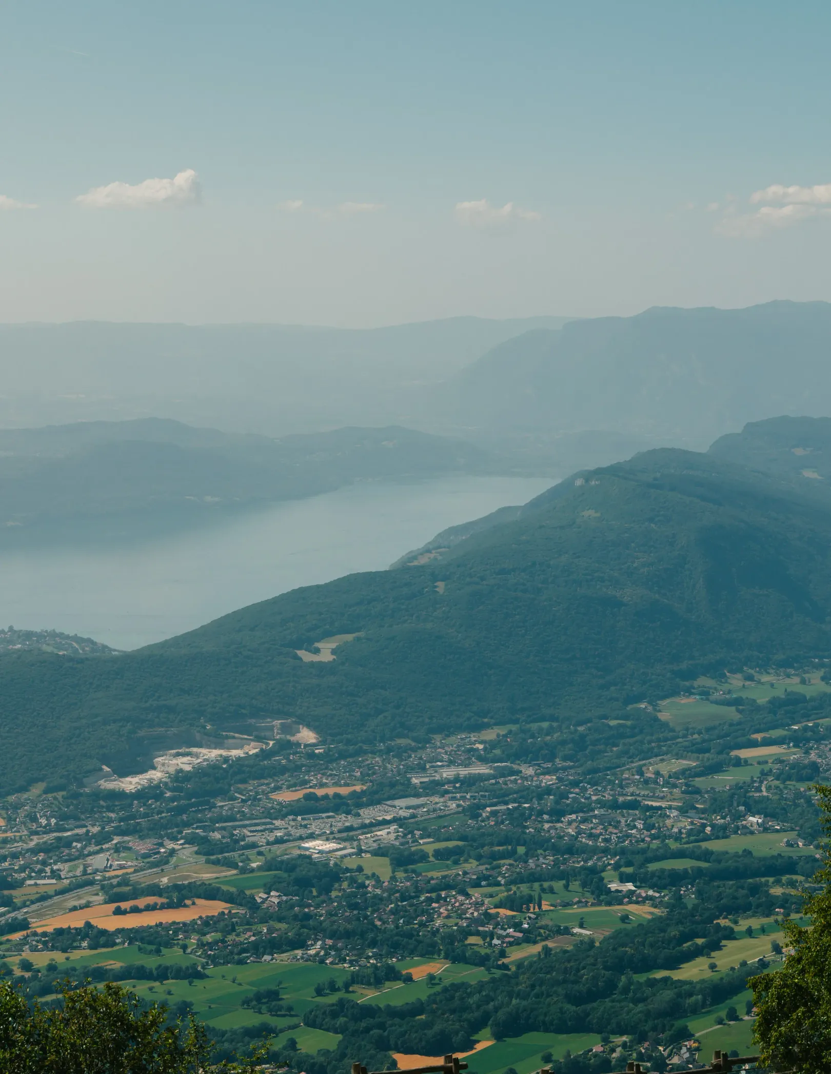 Village entouré de collines verdoyantes avec un lac en arrière-plan sous un ciel légèrement nuageux