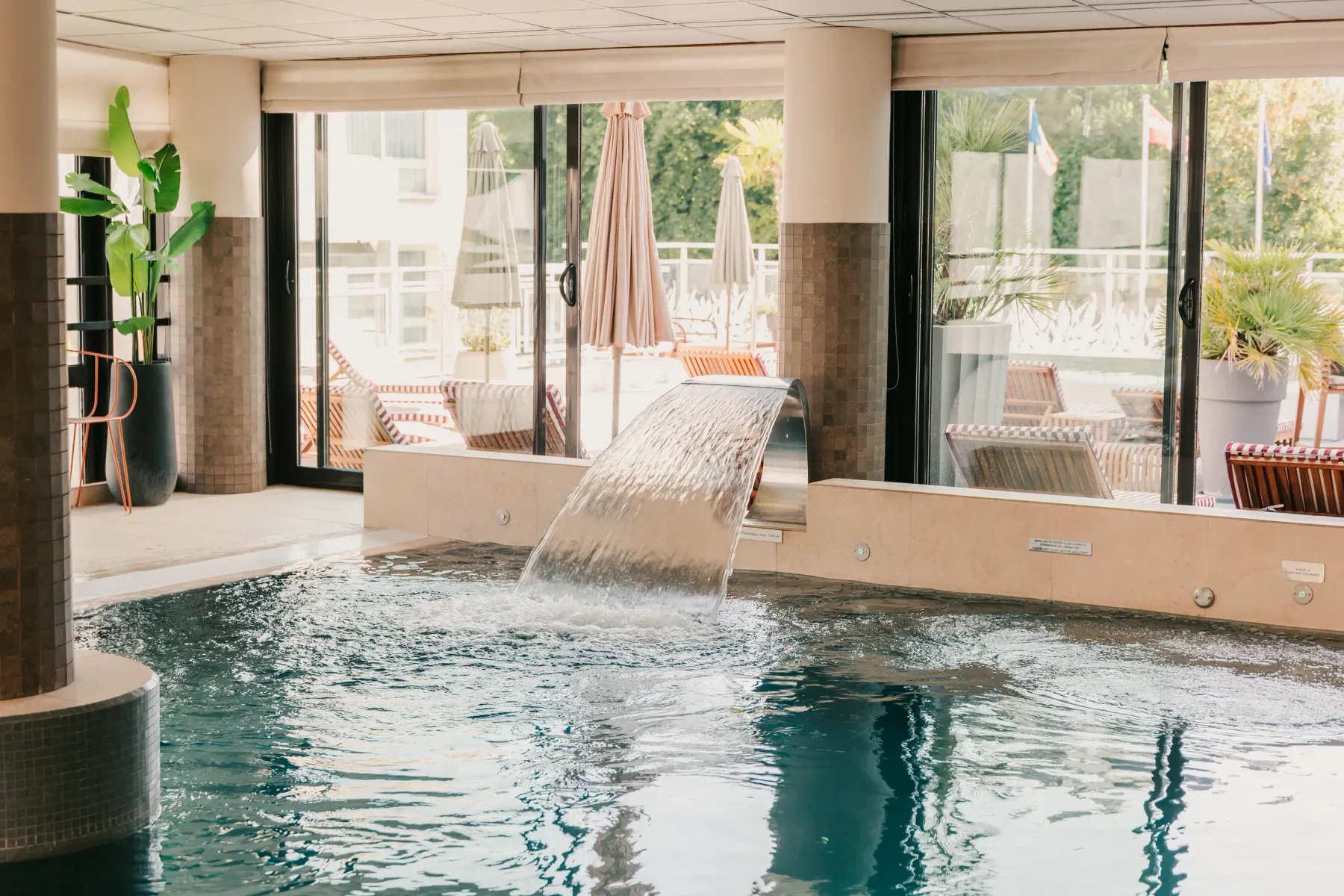 Piscine intérieure avec un petit bassin et une cascade, entourée de grandes baies vitrées ouvrant sur une terrasse avec des chaises longues