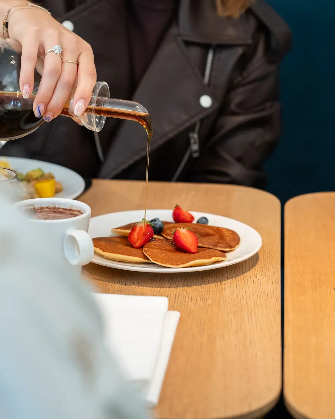 Une personne verse du sirop sur des pancakes garnis de fruits rouges, accompagnés d'une boisson chaude sur une table en bois