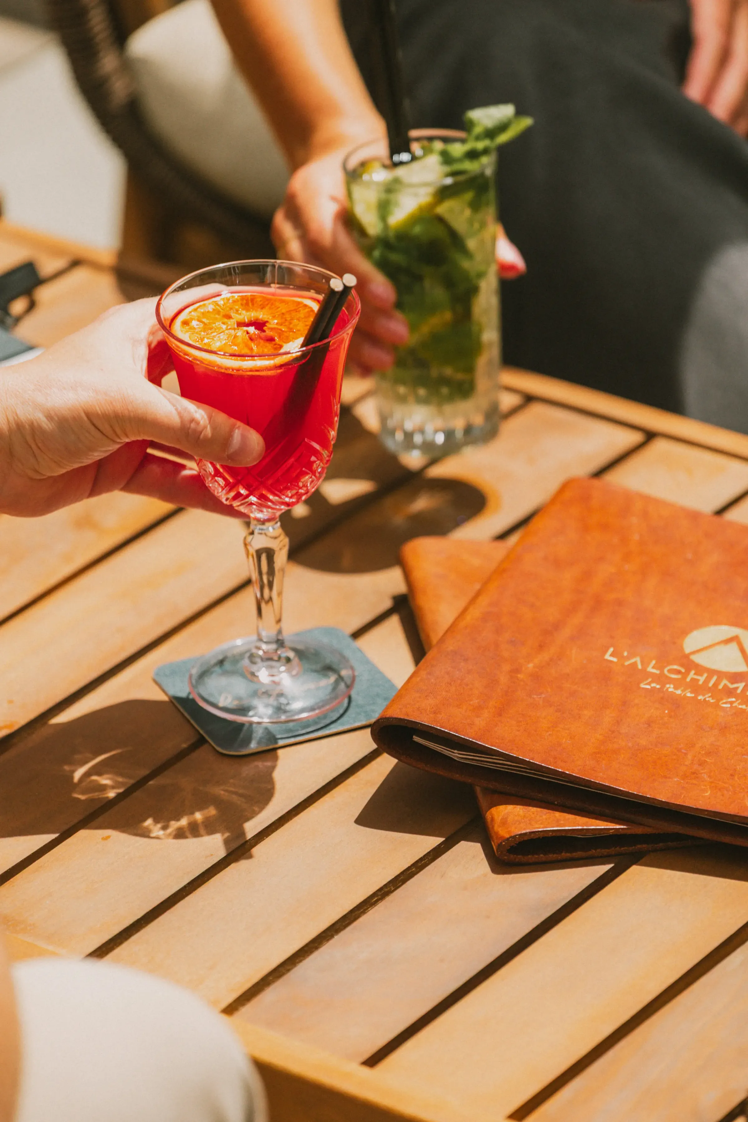 Une main tient un verre de cocktail rouge avec une tranche d'orange, sur une table en bois avec des menus et un verre d'eau